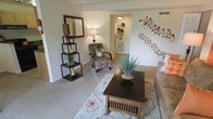 This single-floor residence offers a thoughtfully arranged living space with a welcoming and cozy ambiance. The living room serves as the heart of the home, featuring soft wall-to-wall carpeting underfoot and neutral-toned walls that provide a versatile backdrop for décor. A decorative floral wall mural adds an artistic focal point, giving the room a unique and personalized character. The space is furnished with a patterned accent chair complemented by a bold green throw pillow, a dark wood coffee table, and a sofa, all centered around a floral area rug that ties the room together. A freestanding open shelving unit displays decorative vases and greenery, adding layers of visual interest alongside a warm table lamp that enhances the room's soft lighting. A large framed mirror on the shelving unit reflects light throughout the space, contributing to an airy feel. The layout flows naturally through an open hallway, connecting the living area to the rest of the residence with ease. Visible HVAC vents suggest year-round climate comfort. Overall, the space presents a harmonious blend of functional design and decorative warmth, making it an inviting and livable environment.