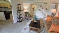 This single-floor residence offers a thoughtfully arranged living space with a welcoming and cozy ambiance. The living room serves as the heart of the home, featuring soft wall-to-wall carpeting underfoot and neutral-toned walls that provide a versatile backdrop for décor. A decorative floral wall mural adds an artistic focal point, giving the room a unique and personalized character. The space is furnished with a patterned accent chair complemented by a bold green throw pillow, a dark wood coffee table, and a sofa, all centered around a floral area rug that ties the room together. A freestanding open shelving unit displays decorative vases and greenery, adding layers of visual interest alongside a warm table lamp that enhances the room's soft lighting. A large framed mirror on the shelving unit reflects light throughout the space, contributing to an airy feel. The layout flows naturally through an open hallway, connecting the living area to the rest of the residence with ease. Visible HVAC vents suggest year-round climate comfort. Overall, the space presents a harmonious blend of functional design and decorative warmth, making it an inviting and livable environment.