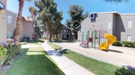 This residential community presents a thoughtfully arranged outdoor environment designed to foster a sense of neighborhood and shared living. The grounds are characterized by expansive, well-maintained lawns that stretch between the buildings, providing open green space for residents to enjoy. Wide concrete walkways wind through the property, connecting various areas of the community and offering convenient pedestrian access throughout.

Mature trees, including tall pines and palms, are distributed across the grounds, providing natural shade and contributing to the established character of the community. The buildings themselves feature a stucco exterior in a neutral gray-brown tone, with select units offering private balconies that overlook the communal spaces below.

A dedicated children's playground is prominently situated within the grounds, equipped with a multi-component play structure featuring stairs, platforms, and a slide. The play area is set on a sand surface, providing a safe and accessible space for younger residents. The overall outdoor environment benefits from abundant natural sunlight, reflecting the warm, sun-drenched climate of the surrounding region, making the common areas inviting throughout the day.