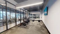 This fitness studio at 16955 W Dixie Hwy in North Miami Beach, FL, is a purpose-built cycling and spin room designed with both functionality and aesthetics in mind. The space is defined by its striking floor-to-ceiling black-framed glass partition walls, which open the room visually to the larger fitness facility beyond while maintaining a focused workout environment. A row of stationary spin bikes is arranged on protective floor mats atop patterned, textured carpet tiles that lend a contemporary feel underfoot. The industrial-inspired ceiling is left exposed, showcasing ductwork and linear LED light fixtures that run the length of the room, providing bright, even illumination throughout. A large flat-screen television is mounted on the white accent wall, offering an engaging visual focal point for cyclists during sessions. Full-length mirrors along one wall further enhance the sense of depth and space. The open layout ensures ease of movement, and the glass walls allow natural light to filter in from the adjacent areas, contributing to an energetic and modern ambiance. The studio's clean, minimalist design and high-quality finishes make it a well-appointed space suited for group cycling classes or individual training sessions.