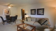 This single-floor space presents a comfortable and traditionally styled living area designed for everyday functionality. The living room serves as the central gathering space, furnished with a plush, beige sectional sofa accented by decorative pillows in neutral tones. A glass-topped, wood-framed coffee table anchors the seating area, complementing the warm, carpeted flooring that runs throughout. A classic black leather armchair adds contrast and additional seating near the corner of the room.

The walls are finished in a clean, off-white tone, providing a neutral backdrop that enhances the room's understated aesthetic. Two framed vintage world map prints are displayed side-by-side along one wall, lending a subtle decorative character to the space. A table lamp with a traditional base provides warm ambient lighting, adding to the inviting atmosphere.

A doorway visible from the living area leads to an adjacent room, suggesting a straightforward, practical layout across the single floor. The overall ambiance is calm and understated, with a classic residential feel supported by consistent neutral tones, soft textures, and timeless furnishing choices throughout the space.