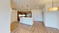 This single-story, one-bedroom, one-bathroom residence offers a thoughtfully designed layout with contemporary finishes throughout. The open-concept living and kitchen area is anchored by light-toned wood-style flooring, recessed lighting, and a ceiling fan, creating a bright and functional space. Large windows allow abundant natural light to fill the living area, which also features a glass-paneled door leading to an outdoor space with tree-lined views.

The kitchen is a standout feature, showcasing rich walnut-toned cabinetry that extends to the ceiling, a white quartz countertop island with a built-in sink, pendant lighting, a textured tile backsplash, and a full suite of stainless steel appliances including a refrigerator, range, and microwave. The bedroom is carpeted for comfort and benefits from dual windows and a ceiling fan. A walk-in closet with dual hanging rods and built-in shelving provides ample storage.

The bathroom features wood-grain cabinetry with a white countertop vanity, wood-style flooring, and a tiled tub-shower combination. The unit's neutral color palette and modern design elements contribute to a clean, contemporary ambiance throughout.