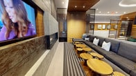This sophisticated single-level lounge space showcases a carefully curated blend of modern materials and warm design elements. The focal point of the room is a striking linear gas fireplace set within a large-format stone tile surround, paired with a wall-mounted flat-screen television — creating a compelling entertainment and relaxation hub. Rich wood paneling lines the walls and ceiling, lending warmth and a refined, contemporary aesthetic throughout the space. A generously proportioned dark charcoal sectional sofa runs along one wall, dressed with an assortment of accent pillows in neutral and teal tones, offering ample seating for guests. In front of the sofa, an arrangement of organic-shaped round wooden side tables clusters atop a boldly striped area rug, adding visual interest and functional surface space. Recessed lighting is integrated into the wood-paneled ceiling, providing a warm, ambient glow that complements the natural tones of the interior. The overall ambiance is polished yet inviting, striking a balance between upscale design and comfortable livability. The cohesive use of stone, wood, and textile materials throughout gives the space a boutique hospitality feel, well-suited for socializing or unwinding in style.