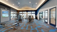 Located at 275 Pine Terrace Drive in Conroe, Texas, this contemporary fitness facility spans a single floor and offers a comprehensive wellness experience. The main exercise room is an expansive, light-filled space anchored by a soaring vaulted ceiling with recessed cove lighting and dual ceiling fans. Bold navy blue geometric accents complement the eye-catching blue-and-gray patterned carpet throughout. Floor-to-ceiling windows along multiple walls flood the room with natural light and provide views of the surrounding community. The space is equipped with a full array of strength-training machines, free weights, treadmills, ellipticals, stair climbers, and large-screen televisions. A secondary exercise room features a striking blue-and-white striped accent wall, hardwood flooring, wall-to-wall mirrors, and a dedicated row of smart spin bikes with individual display screens. A strength area with a power rack and bench is also available, accompanied by motivational wall signage. The facility also includes a vibrant children's lounge featuring colorful modular seating, built-in toy and book shelving with accent lighting, playful wall murals, and large windows overlooking the pool area — creating a welcoming space for younger visitors.