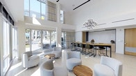 This contemporary communal space is defined by its striking double-height ceilings, which lend an airy, open feel throughout. Floor-to-ceiling windows and glass doors flood the interior with natural light while offering direct views and access to an outdoor area, likely a pool or courtyard. Plaid-patterned drapery adds a subtle textural contrast to the otherwise crisp, white palette.

The lounge is thoughtfully divided into two functional zones. A casual seating area features rounded, light gray upholstered chairs arranged around small circular wood-topped tables, creating an inviting space for relaxed gatherings. Adjacent to this, a bar-height communal table with a warm wood surface is flanked by dark metal stools, serving as an ideal workspace or social hub. Above this area, a geometric, multi-arm chandelier serves as a bold focal point.

Light hardwood flooring runs throughout, complementing the neutral tones of the furniture and walls. Track lighting on the ceiling supplements the abundant daylight. The overall ambiance is clean, modern, and welcoming, balancing functionality with refined aesthetic detail.