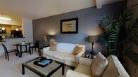 This single-floor space offers a well-appointed interior designed with a contemporary aesthetic and a carefully curated color palette. The living area serves as the centerpiece of the space, showcasing a striking slate-blue accent wall that contrasts elegantly with the lighter surrounding walls and ceiling. A plush cream sofa with coordinating accent cushions anchors the seating arrangement, complemented by a matching armchair and a sleek glass-top coffee table with a dark frame. Stylish table lamps with dark bases provide warm, ambient lighting throughout the room, enhancing the inviting atmosphere. A framed wall mirror adds depth and visual interest to the accent wall, while a tall indoor plant introduces a refreshing natural element to the interior. A compact dining or work area with a dark table and chairs is visible in the background, adding functional versatility to the layout. The overall design balances modern sophistication with comfort, featuring consistent use of neutral and muted tones, dark accents, and carefully chosen furnishings that contribute to a cohesive and polished living environment.