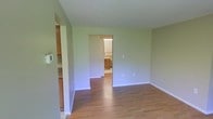 This single-floor residential space offers a clean, functional layout designed for comfortable everyday living. The interior is finished with warm-toned hardwood flooring that extends throughout the main living area, adding a sense of continuity and warmth to the space. The walls are painted in a soft, muted green that creates a calm and neutral ambiance, complementing the natural tones of the flooring.

The main living area is open and well-proportioned, with smooth white ceilings and clean white baseboards that frame the space neatly. A doorway on one side provides direct access to a bathroom, which features a vanity with wooden cabinetry and appears bright and functional. To the opposite side, a partial view reveals an adjacent kitchen area with wooden cabinetry and countertops, offering convenient connectivity between living and cooking spaces.

Natural light filters into the space, enhancing its airy and uncluttered feel. The layout flows naturally between rooms, making it easy to navigate. Overall, this space presents a modest yet well-maintained interior with consistent finishes throughout, making it a practical and inviting single-floor residence.