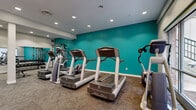 Located at 205 Vine St in Columbus, Ohio, this fitness center offers a well-equipped exercise space designed to support a comprehensive workout routine. The room is finished with a bold teal accent wall that adds energy and visual interest, complemented by neutral gray carpet flooring and crisp white walls throughout. Recessed ceiling lighting ensures the space is consistently bright and inviting.

The cardio section is a highlight of the facility, featuring multiple Life Fitness treadmills, elliptical machines, and a stair climber, all arranged to allow comfortable use. Large floor-to-ceiling windows along one wall invite natural light and provide outdoor views, enhancing the workout atmosphere. A Life Fitness functional training cable cross machine serves as a centerpiece for strength training, while a mirrored wall section reflects the space and visually expands the room.

A dedicated free weights area includes a dumbbell rack and exercise ball storage, supporting a variety of strength and flexibility routines. Wall-mounted flat-screen televisions and vibrant orange storage cabinets add functional and aesthetic touches. A branded entrance mat near the glass entry door adds a welcoming, polished finish to this comprehensively appointed fitness center.