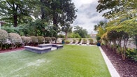 Located at 700 Woodrow St in Columbia, South Carolina, this thoughtfully designed outdoor space offers a refined and inviting environment ideal for relaxation and entertaining. The grounds feature expansive, well-maintained artificial turf that provides a clean, verdant centerpiece to the entire area.

A tiled patio area serves as a functional outdoor living zone, equipped with a cast-iron dining table and chairs shaded by a large umbrella, alongside a full-sized gas grill — perfect for al fresco dining. A patio heater ensures year-round usability of the space. A separate lounge area along one side of the grounds offers multiple chaise lounges arranged for sunbathing or leisure.

A raised stone feature — likely a fire pit or water feature — adds architectural interest and serves as a focal point within the landscape. Lush, layered plantings line the perimeter, including Japanese maples, magnolias, palms, ornamental shrubs, and vibrant potted plants, all set against a clean stucco privacy wall that encloses the space. Mature trees provide natural shade and a sense of seclusion.

The overall ambiance is that of a serene, resort-style retreat, combining practical outdoor amenities with carefully curated landscaping in the heart of Columbia's Elmwood Park neighborhood.