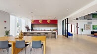 This generously sized resident lounge in Miami, FL is a thoughtfully designed communal space that seamlessly blends social, recreational, and relaxation areas into one cohesive environment. The space is defined by its bold architectural details, including striking red coffered and textured ceiling panels that contrast dramatically against crisp white walls, creating a dynamic visual identity throughout.

The main living areas feature large-format porcelain tile flooring, floor-to-ceiling glass walls and sliding doors that flood the interiors with natural light and offer open views of lush tropical greenery outside. A dramatic striped ceiling element in the entry zone adds a graphic, contemporary touch. Seating areas are furnished with a mix of leather sofas, modular white couches, and boldly colored accent chairs, anchored by herringbone-patterned area rugs.

A dedicated lounge zone includes a wall-mounted TV and comfortable seating for media viewing, while a billiards table provides additional recreation. Private dining-style booths with deep red upholstery and pendant lighting offer intimate gathering spots. A communal kitchen and bar area showcases a marble island, rose-toned cabinetry, and sculptural copper pendant lights. A co-working zone features a long wood table with blue task chairs, complemented by pod-style lounge seating for flexible use throughout the day.