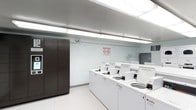 This shared laundry facility is a single-floor space designed with functionality and cleanliness in mind. The room features a row of top-load washing machines lined along one wall, numbered for easy identification and equipped with individual coin or card payment mechanisms. The machines are arranged in a neat, accessible configuration that maximizes the usable floor space. A tall, dark-toned multi-door storage locker unit stands prominently along the adjacent wall, offering secure storage options for residents or users of the facility. The space is illuminated by bright fluorescent strip lighting mounted to the ceiling, ensuring excellent visibility throughout. The walls are finished in clean white painted cinder block, contributing to the room's crisp, utilitarian aesthetic. The floor features a white textured tile that is both practical and easy to maintain. A wall-mounted utility area near the back of the room includes a hanging rod and shelf, providing additional convenience for users. Safety and instructional notices are posted on the wall, reflecting a well-managed and organized facility. Overall, the space is efficiently laid out, well-maintained, and purpose-built to serve the laundry needs of its users.