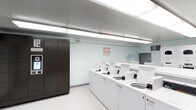 This shared laundry facility is a single-floor space designed with functionality and cleanliness in mind. The room features a row of top-load washing machines lined along one wall, numbered for easy identification and equipped with individual coin or card payment mechanisms. The machines are arranged in a neat, accessible configuration that maximizes the usable floor space. A tall, dark-toned multi-door storage locker unit stands prominently along the adjacent wall, offering secure storage options for residents or users of the facility. The space is illuminated by bright fluorescent strip lighting mounted to the ceiling, ensuring excellent visibility throughout. The walls are finished in clean white painted cinder block, contributing to the room's crisp, utilitarian aesthetic. The floor features a white textured tile that is both practical and easy to maintain. A wall-mounted utility area near the back of the room includes a hanging rod and shelf, providing additional convenience for users. Safety and instructional notices are posted on the wall, reflecting a well-managed and organized facility. Overall, the space is efficiently laid out, well-maintained, and purpose-built to serve the laundry needs of its users.