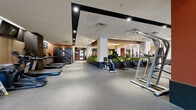 This well-appointed fitness center offers a comprehensive workout environment spread across a single floor, thoughtfully divided into multiple distinct training zones. The cardio area features a generous lineup of treadmills, ellipticals, stair climbers, stationary bikes, and air bikes, all positioned alongside flat-screen monitors for entertainment during workouts. Warm wood-panel accents along the walls, bold terracotta and dark charcoal tones, and recessed lighting create a modern and energizing atmosphere throughout. A dedicated strength training zone is equipped with cable machines, squat racks, adjustable benches, and free weights, while large mirrors amplify the sense of space. A functional training area provides battle ropes, kettlebells, medicine balls, TRX suspension trainers, and a pull-up rig, set against natural light from street-facing windows and wrapped in natural wood paneling. One of the facility's most distinctive features is the central planter installation, filled with lush tropical greenery that softens the industrial aesthetic and contributes to a refreshing, biophilic ambiance. A separate open-floor studio space with light wood flooring and parallel bars offers room for stretching, mobility work, or group training. The rubber flooring throughout ensures safety and comfort across all zones.