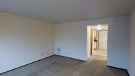 This two-floor residential space presents a straightforward and functional layout designed for comfortable everyday living. The main living area is finished with wall-to-wall carpeting in a neutral tone, contributing to a quiet and cozy atmosphere. Smooth white walls and a flat ceiling keep the space feeling clean and uncluttered, while a floor-level heating vent ensures year-round climate comfort. An open doorway connects the living area directly to the kitchen, which features light-colored cabinetry and tile or vinyl flooring, offering a seamless flow between the primary gathering space and the cooking area. The corridor connecting the two rooms is lined with warm wood-toned trim, adding a subtle accent to the otherwise minimal interior palette. Recessed and flush-mount ceiling lighting provides consistent illumination throughout. The overall design prioritizes practicality and ease of use, with a neutral color scheme throughout that offers a versatile backdrop for a variety of furnishing styles. Spanning two floors, the space provides ample room for both living and private quarters, making it a well-organized residence suited for individuals or small households seeking a no-frills, functional home environment.