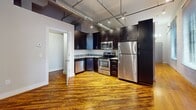 Unit ITL 106 is a 520-square-foot, single-floor, 2-bedroom loft-style apartment that blends industrial aesthetics with functional living. The open-concept layout is anchored by rich, warm hardwood floors that run throughout the entire unit, adding character and continuity to the space.

The kitchen is a standout feature, outfitted with dark espresso cabinetry, granite countertops, and a full suite of stainless steel appliances including a refrigerator, range, microwave, and dishwasher. Exposed ductwork and industrial-style track lighting on the ceiling reinforce the loft aesthetic and provide practical illumination.

The two bedrooms are simply appointed with neutral gray walls, hardwood floors, and access to closet spaces. One bedroom features exposed white-painted brick walls and a large window fitted with blinds, allowing natural light to filter in while maintaining privacy. The other bedroom benefits from similar industrial ceiling details, maintaining design consistency throughout the unit.

The overall ambiance of ITL 106 is urban and industrial, characterized by high ceilings with exposed mechanical systems, original hardwood floors, and raw architectural details. This unit is well-suited for those who appreciate a modern loft aesthetic within a compact and efficient floor plan.
