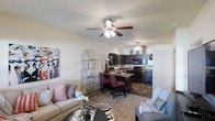 This single-floor residence showcases an open-concept design that integrates the living, dining, and kitchen areas into one cohesive and functional space. The living room is warm and inviting, featuring plush carpeted flooring, a ceiling fan with integrated lighting, and ample room for comfortable seating arrangements. Decorative touches, including wall art and an open display shelving unit, add personality and character to the space.

The kitchen is well-appointed with rich, dark cabinetry that provides generous storage, black appliances including a refrigerator, stove, and microwave, and a kitchen island or breakfast bar with seating for two or three, making it ideal for casual dining or entertaining. The kitchen flows naturally into the dining area, creating a smooth transition between cooking and gathering spaces.

Throughout the space, neutral wall tones complement the darker cabinetry and furnishings, contributing to a balanced and cohesive aesthetic. Recessed lighting in the kitchen area, combined with ceiling fans in both the living and kitchen zones, ensures the space remains well-lit and comfortable. The overall ambiance is casual yet stylish, offering a practical and welcoming environment suited for everyday living.
