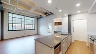 This single-floor loft unit blends industrial character with modern finishes throughout. Soaring ceilings with exposed ductwork, painted structural beams, and original brick walls speak to the building's industrial heritage, while wide-plank hardwood flooring and crisp white walls create a clean, contemporary contrast.

The open-concept living and kitchen area is a standout feature. A generously sized central island with a dark quartz countertop, integrated sink, and built-in appliances anchors the kitchen, which is accented by a distinctive powder-blue base and white upper cabinetry. A stainless steel range hood, full-size refrigerator, and a built-in wine rack add both function and character. Recessed lighting illuminates the space alongside abundant natural light pouring through oversized, steel-framed factory windows.

The living area is expansive and open, flowing seamlessly from the kitchen. A dedicated laundry closet with a stacked washer and dryer unit is conveniently tucked off the kitchen corridor. The bedroom features large windows with exterior views and warm hardwood floors.

The bathroom is well-appointed with large-format tile flooring, full-height tile surrounding a soaking tub and shower combination, a warm wood vanity with a white countertop, and open shelving for additional storage. The overall ambiance is airy, bright, and distinctly urban.