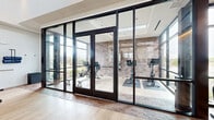 This professionally designed fitness center offers a generous, open workout environment thoughtfully arranged across a single floor. The space makes an immediate impression with its soaring vaulted ceilings accented by warm wood paneling, which contrast beautifully against crisp white walls and light hardwood flooring throughout. A striking floor-to-ceiling glass wall system with sleek black-framed doors and transoms divides a dedicated cycling and cardio alcove from the main workout floor, flooding both areas with abundant natural light and providing clear sightlines across the entire facility. Exposed brick detailing adds a textural, industrial-chic warmth to the cycling studio, while large windows along the perimeter frame outdoor views and enhance the airy atmosphere. The main floor is comprehensively equipped with a full array of Precor strength machines, cable systems, a squat rack, and an organized free-weight station featuring a complete dumbbell rack alongside an interactive mirror display. The cardio zone houses multiple treadmills, elliptical trainers, and stationary bikes. Full-length mirrors extend the visual depth of the space. The combination of high-end equipment, sophisticated architectural finishes, and exceptional natural light creates a bright, motivating, and premium fitness environment.