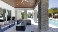 This covered outdoor living space offers a seamless blend of natural materials and modern design, creating a comfortable and visually striking environment. Stacked stone columns define the perimeter of the space, lending a sense of permanence and architectural character while framing open views of the surrounding pool area and manicured landscaping beyond.

The ceiling is finished with warm wood paneling, adding a rich, organic contrast to the stone elements and contributing to an inviting atmosphere. Recessed lighting and a large ceiling fan are integrated into the ceiling, ensuring comfort and functionality throughout the day and into the evening.

The floor is laid with large-format light stone or porcelain tiles, which complement the neutral tones of the columns and reflect natural light effectively. The outdoor lounge area is furnished with dark wicker seating accented by bold, multicolored striped cushions, centered around a glass-top coffee table.

Beyond the columns, views open to a pool deck lined with lounge chairs, tall hedges, and lush greenery, reinforcing the private and resort-like character of the space. This area functions as a transitional zone between interior and exterior living.