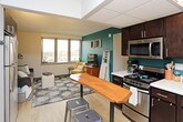 This single-floor unit offers a thoughtfully arranged layout encompassing a living area, kitchen, bedroom, and full bathroom. The living room is a welcoming space highlighted by a wide bank of windows that frame outdoor views and fill the room with natural light. A bold teal accent wall adds a vibrant design element, complementing the light hardwood-style flooring that runs throughout the main living and kitchen areas.

The kitchen is efficiently designed, featuring dark espresso cabinetry, stainless steel appliances including a range and microwave, light countertops, and a teal tile backsplash that ties in with the living area's color palette. A bistro-style high table with metal stools sits adjacent to the kitchen, providing a casual dining option near the entry.

The bedroom is carpeted for comfort and benefits from natural light through horizontal-blind windows. It includes space for a desk and chair, offering functional utility alongside the sleeping area. A full-length ornate mirror adds a decorative touch. The bathroom features light wood-style vinyl flooring, a dark vanity with an integrated sink, and a shower-tub combination with a curtain enclosure, completing this practical and cohesive living space.