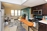This single-floor unit offers a thoughtfully arranged layout encompassing a living area, kitchen, bedroom, and full bathroom. The living room is a welcoming space highlighted by a wide bank of windows that frame outdoor views and fill the room with natural light. A bold teal accent wall adds a vibrant design element, complementing the light hardwood-style flooring that runs throughout the main living and kitchen areas.

The kitchen is efficiently designed, featuring dark espresso cabinetry, stainless steel appliances including a range and microwave, light countertops, and a teal tile backsplash that ties in with the living area's color palette. A bistro-style high table with metal stools sits adjacent to the kitchen, providing a casual dining option near the entry.

The bedroom is carpeted for comfort and benefits from natural light through horizontal-blind windows. It includes space for a desk and chair, offering functional utility alongside the sleeping area. A full-length ornate mirror adds a decorative touch. The bathroom features light wood-style vinyl flooring, a dark vanity with an integrated sink, and a shower-tub combination with a curtain enclosure, completing this practical and cohesive living space.