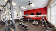 Located at 1450 Bluewater Way in Charleston, SC, this fitness center is a well-equipped, single-room facility designed to support a comprehensive workout routine. The space is defined by a bold design aesthetic, featuring a striking red accent wall accented with white horizontal stripes, providing an energizing atmosphere for exercise.

The cardio zone lines the accent wall and includes multiple treadmills, an elliptical trainer, a stationary bike, and a rowing machine, each equipped with individual digital display consoles. Three wall-mounted flat-screen televisions are positioned above the cardio equipment, allowing users to stay entertained during their sessions.

The strength training area occupies the opposite side of the room and is anchored by a multi-functional cable machine and Smith machine, along with a fully stocked dumbbell rack and adjustable weight benches. Full-wall mirrors along one side of the room visually expand the space and allow users to monitor their form during lifts.

Durable rubber flooring covers the entire room, providing both safety and sound dampening. Ceiling fans with integrated lighting fixtures and fluorescent overhead lighting ensure the space remains well-lit and comfortable. Natural light filters in through windows fitted with blinds. A glass-paneled door offers a direct exit, adding to the room's accessibility and ventilation.