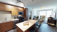 This thoughtfully designed single-floor residence showcases a seamless open-concept layout that connects the kitchen, dining, and living areas into one cohesive and inviting space. The kitchen is a standout feature, appointed with warm wood cabinetry, stainless steel appliances including a gas range, microwave, and dishwasher, and a generous center island with a clean white countertop that doubles as a breakfast bar with seating. Stylish pendant lighting hangs above the island, complementing the sleek track lighting overhead and adding a modern, curated aesthetic. The adjacent dining area accommodates a full table and chairs, flowing naturally into the living room beyond. Large windows flood the living area with natural light, creating a bright and airy atmosphere throughout. The flooring transitions seamlessly across the open plan, unifying the space visually. Neutral tones are balanced by warm wood accents throughout the cabinetry and furnishings, lending a contemporary yet welcoming ambiance. A hallway visible from the main area suggests additional private rooms beyond, completing this well-appointed and functional single-floor residence.