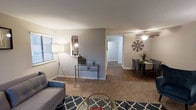 This single-floor residence offers two bedrooms and one and a half bathrooms across a thoughtfully arranged floor plan. The open living and dining area forms the heart of the home, featuring warm hardwood-style flooring in the dining zone that transitions seamlessly into plush carpeting in the living room. The living area is generously sized, providing ample space for comfortable furnishings, and is brightened by natural light filtering through large windows dressed with horizontal blinds. A neutral, two-tone color palette of soft white and cool gray across the walls lends the space a clean, modern aesthetic. A decorative accent wall in the living area adds visual depth and character. The dining area is anchored by a ceiling fan with a light fixture, offering both functionality and style, and comfortably accommodates a full dining set. A central hallway connects the main living spaces to the bedrooms, ensuring a logical and private layout. The half bathroom adds convenience for guests, while the full bathroom serves the bedrooms. Overall, this residence presents a practical and inviting atmosphere, well-suited for everyday living with a balance of open communal areas and private sleeping quarters.
