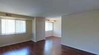 This single-floor residence presents a welcoming and straightforward layout designed for comfortable everyday living. The main living area is defined by rich, dark hardwood flooring that extends throughout, providing warmth and visual depth to the space. Neutral, light-toned walls create a clean and versatile backdrop, allowing for a variety of interior design possibilities. Natural light filters in through vertical-blind-covered windows, brightening the interior and contributing to an airy ambiance. A flush-mount ceiling light fixture provides additional illumination, ensuring the space remains well-lit at all hours. The room opens into a secondary area — likely a bedroom or flex space — separated by a standard interior door, maintaining a sense of defined zones within the open flow of the unit. Floor-level air vents are integrated into the walls, supporting climate control throughout. The white baseboards and trim offer a crisp contrast against the darker flooring, adding a subtle touch of architectural polish. Overall, the space presents a clean, move-in-ready environment with functional design elements and a neutral palette that suits a wide range of uses.