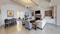 This single-floor space offers a thoughtfully designed open layout that seamlessly connects key functional areas. The expansive living room serves as the centerpiece, featuring soaring vaulted ceilings with exposed beams, a striking stacked-stone fireplace topped with a wall-mounted television, and polished concrete flooring throughout. An elegant chandelier and recessed lighting provide layered illumination, while large windows and glass-paneled entry doors allow generous natural light to fill the room. The living area accommodates comfortable seating arrangements, lending the space a welcoming, lounge-like atmosphere.

Adjacent to the living room, the kitchen is well-equipped and efficiently organized, showcasing rich dark cabinetry with brushed hardware, light granite countertops, a subway tile backsplash, and a full suite of stainless steel appliances including a gas range with a wall-mounted hood vent, dishwasher, and a large French door refrigerator. Pendant lighting and a window above the sink enhance both function and ambiance. A dedicated dining area nearby features clean white walls and ample room for a dining table, completing this versatile, communal-feeling space.