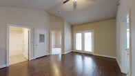 This single-story space is defined by its striking vaulted ceilings, which lend an airy, expansive quality to the main living area. The soaring ceiling height is complemented by a ceiling fan, ensuring both comfort and visual appeal. Underfoot, rich dark hardwood flooring runs throughout, adding warmth and a polished finish to the space.

The main living area is bathed in natural light, courtesy of a set of classic French doors fitted with blinds, offering both brightness and privacy control. These doors likely provide access to an outdoor area, further enhancing the sense of openness. The neutral, warm beige wall tones create a versatile backdrop that works well with a variety of interior styles.

A doorway visible from the main living area leads to an additional room, suggesting a well-connected floor plan. A secondary door, likely a utility or exterior access point, is also present and features a ventilation panel. The overall ambiance of the space is clean, open, and inviting, with its combination of high ceilings, natural light, and quality flooring creating a welcoming environment suited to a range of uses.