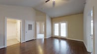 This single-story space is defined by its striking vaulted ceilings, which lend an airy, expansive quality to the main living area. The soaring ceiling height is complemented by a ceiling fan, ensuring both comfort and visual appeal. Underfoot, rich dark hardwood flooring runs throughout, adding warmth and a polished finish to the space.

The main living area is bathed in natural light, courtesy of a set of classic French doors fitted with blinds, offering both brightness and privacy control. These doors likely provide access to an outdoor area, further enhancing the sense of openness. The neutral, warm beige wall tones create a versatile backdrop that works well with a variety of interior styles.

A doorway visible from the main living area leads to an additional room, suggesting a well-connected floor plan. A secondary door, likely a utility or exterior access point, is also present and features a ventilation panel. The overall ambiance of the space is clean, open, and inviting, with its combination of high ceilings, natural light, and quality flooring creating a welcoming environment suited to a range of uses.
