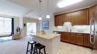 This single-floor residence in Mill offers a thoughtfully designed open-concept layout that connects the kitchen, dining, and living areas in a seamless, functional flow. The kitchen is a standout feature, showcasing warm honey-toned cabinetry with ample storage, a classic subway tile backsplash, and light granite countertops that extend across a spacious central island. The island is complemented by stylish pendant lighting and black bar stools, making it an ideal spot for casual dining or entertaining. Stainless steel appliances and an undermount sink add a polished, modern touch. The open layout transitions naturally into the living and dining areas, which are bathed in natural light streaming through large sliding glass doors. These doors provide direct access to an outdoor patio or balcony, offering pleasant views of the surrounding exterior. The flooring throughout features a combination of light tile in the kitchen area and plush carpeting in the living spaces, providing both practicality and comfort. Neutral wall tones and recessed lighting contribute to a bright, airy ambiance throughout. The overall design balances everyday functionality with an inviting, comfortable aesthetic well-suited for modern living.