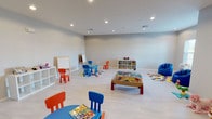 This generously sized children's playroom is thoughtfully designed to provide an engaging and organized environment for young children. The space features light-toned, smooth flooring throughout that is both easy to clean and comfortable for play. Neutral gray walls and recessed ceiling lighting create a bright, welcoming ambiance while keeping the focus on the colorful furnishings and activity areas within.

The room is divided into several distinct play zones. A child-sized activity table with vibrant orange and blue chairs serves as a central hub for arts, crafts, and games, while a dedicated art corner features both a whiteboard and a chalkboard easel for creative expression. A wooden train table provides an interactive focal point at the center of the space, surrounded by colorful patchwork area rugs that define individual play areas.

White cube shelving units line portions of the walls, offering organized storage for toys, books, and collectibles. A play kitchen station and a vanity set add imaginative role-play opportunities, while a plush blue bean bag chair offers a cozy reading or relaxation nook. A window with blinds allows natural light to filter into one section of the room.

An adjacent doorway connects to a separate recreation area, visible in the background, indicating that this playroom is part of a larger entertainment-focused floor plan. Overall, the space is clean, functional, and purposefully designed for children's enrichment and enjoyment.