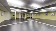 This purpose-built studio space is designed to support movement, dance, or fitness activities across a single open floor. The studio's most defining feature is its expansive floor-to-ceiling mirror panels, which line the walls and visually amplify the room, creating a sense of generous, open space ideal for classes or individual practice. The smooth, grey hardwood-style flooring provides a clean and resilient surface well-suited for a variety of activities. Overhead, a grid of fluorescent strip lighting integrated into a drop ceiling ensures the entire space is evenly and brightly illuminated at all times. Structural white columns punctuate the room while painted yellow-green accent walls lend a warm, energizing ambiance. A wooden bench along one wall offers a convenient area for resting or changing footwear, and framed artwork adds a welcoming touch to the environment. The studio is equipped with a camera or lighting tripod setup near the entrance, suggesting capability for recording or livestreaming sessions. Emergency exit signage is clearly visible, and the layout provides unobstructed open floor space, making it highly functional and accessible for instructors and participants alike.