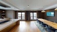 This thoughtfully designed single-floor space seamlessly blends function and contemporary elegance. The open-concept layout connects a professionally appointed kitchen with a formal dining and meeting area, creating a versatile environment suited for gatherings, events, or professional meetings.

The kitchen is a standout feature, outfitted with sleek taupe cabinetry accented by brass hardware, a striking marble-topped countertop with a brass bridge faucet, and a gas range. Open corner shelving adds a curated display element, while large black-framed sliding glass doors flood the space with natural light and provide direct access to an outdoor terrace — ideal for extended entertaining.

The dining and meeting area is anchored by a substantial natural wood table surrounded by upholstered navy barrel chairs set atop an ornate area rug. Two wall-mounted displays serve as functional presentation screens, while a trio of white dome pendant lights with gold interiors cast warm, focused illumination. The deep charcoal accent walls throughout lend a moody, refined atmosphere that contrasts beautifully with the light hardwood floors and white tray ceiling details.

Recessed ceiling speakers and integrated smart controls further enhance the space's utility, making it equally suited for formal presentations or sophisticated social occasions. The combination of natural materials, brass accents, and modern finishes creates a cohesive, upscale aesthetic throughout.