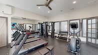 Located at 2745 Campus Pointe Cir in Gainesville, Georgia, this fitness center is a well-equipped, single-floor facility designed to support a comprehensive workout experience. The space is accessed through charming multi-pane glass French doors that open from a wood-floored entryway, setting a clean and modern tone upon arrival. Inside, the main workout area features durable rubber flooring throughout, providing both safety and comfort during exercise. A ceiling fan overhead aids in air circulation, complemented by recessed lighting that keeps the space bright and functional. The cardio section is outfitted with multiple Star Trac treadmills arranged in a row, alongside an elliptical trainer and a stationary cycle, catering to a range of cardiovascular preferences. A multi-functional cable strength machine serves as the centerpiece of the strength training area, flanked by a well-stocked dumbbell rack offering a variety of weight options. A flat workout bench and a Titan plyo box round out the functional training equipment. Full-length mirrors line one wall, visually expanding the space and allowing users to monitor their form. A wall-mounted television adds an entertainment element for those who enjoy media during their workouts. Natural light filters in through windows along one wall, contributing to an open and energizing atmosphere throughout the facility.