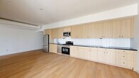Located in the vibrant East Hollywood neighborhood of Los Angeles, this modern 2-bedroom, 2-bathroom single-floor residence offers a thoughtfully designed open-concept layout. Light-toned hardwood floors run throughout the space, creating a warm and cohesive aesthetic. The kitchen is a standout feature, showcasing an extended run of light wood cabinetry — both upper and lower — paired with dark countertops and a clean white subway tile backsplash. Stainless steel appliances, including a gas range, microwave, and refrigerator, complete the functional and contemporary kitchen. Exposed ductwork along the ceiling adds an industrial touch to the bright, airy living area. The bedrooms are bright and minimal, with clean white walls; the primary bedroom features a large window that frames views of the surrounding Los Angeles neighborhood and fills the room with natural light. Both bathrooms are finished with striking floor-to-ceiling green glazed tile in the walk-in showers, terrazzo flooring, and natural wood vanities with matte black hardware, lending a spa-like quality. Hallways connect the space efficiently, and the overall design reflects a clean, modern aesthetic well-suited to urban Los Angeles living.