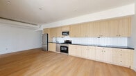 Located in the vibrant East Hollywood neighborhood of Los Angeles, this modern 2-bedroom, 2-bathroom single-floor residence offers a thoughtfully designed open-concept layout. Light-toned hardwood floors run throughout the space, creating a warm and cohesive aesthetic. The kitchen is a standout feature, showcasing an extended run of light wood cabinetry — both upper and lower — paired with dark countertops and a clean white subway tile backsplash. Stainless steel appliances, including a gas range, microwave, and refrigerator, complete the functional and contemporary kitchen. Exposed ductwork along the ceiling adds an industrial touch to the bright, airy living area. The bedrooms are bright and minimal, with clean white walls; the primary bedroom features a large window that frames views of the surrounding Los Angeles neighborhood and fills the room with natural light. Both bathrooms are finished with striking floor-to-ceiling green glazed tile in the walk-in showers, terrazzo flooring, and natural wood vanities with matte black hardware, lending a spa-like quality. Hallways connect the space efficiently, and the overall design reflects a clean, modern aesthetic well-suited to urban Los Angeles living.