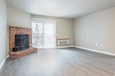 This single-floor unit is designed with a practical, open layout that connects the kitchen and living areas seamlessly. The kitchen is equipped with white cabinetry featuring warm wood-tone accents, a double stainless steel sink, white electric range, built-in microwave, and a full-size refrigerator. A peninsula counter extends into the living space, providing additional prep and seating opportunities while maintaining an open, connected feel throughout.

The living area is a generously sized, light-filled space highlighted by a corner brick fireplace, adding a classic architectural focal point. A sliding glass door with vertical blinds allows natural light to pour in and provides outdoor access. Light-toned laminate flooring runs consistently throughout the unit, contributing to a cohesive, clean aesthetic.

The bedroom features a notable vaulted ceiling that adds an airy, spacious quality to the room. Two side-by-side bifold closets offer ample storage, and a window provides natural light. An additional single closet with a hanging rod is also present elsewhere in the unit. The bathroom includes a white vanity with a round mirror, a full-size tub and shower combination, and neutral, understated finishes throughout.