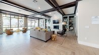 This single-floor community clubhouse is thoughtfully designed to offer a sophisticated and comfortable gathering space. The grand entrance foyer sets an elegant tone with polished marble flooring, soaring double-height ceilings, and tall glass doors that flood the space with natural light and frame views of swaying palm trees outside.

Beyond the foyer, the main lounge area is anchored by a wall-mounted fireplace with a sleek tile surround and a large flat-screen TV above. Plush seating, including a generous sectional sofa and accent chairs, is arranged to encourage conversation and relaxation. The striking herringbone-patterned wood-look tile flooring extends throughout, complemented by dramatic dark wood coffered ceilings that add architectural depth and character.

A well-appointed kitchenette features sage-green cabinetry, subway tile backsplash, dark countertops, a beverage refrigerator, and a coffee station, making it ideal for hosting and social gatherings. Adjacent to the kitchenette, a billiards area with cue storage adds a recreational dimension to the space.

A bright co-working nook lined with expansive floor-to-ceiling windows offers counter-height seating and unobstructed views of the lush, palm-lined outdoor courtyard. Patterned drapery and recessed lighting throughout contribute to a warm yet refined ambiance, making this clubhouse a versatile and welcoming destination.