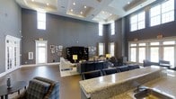 The Club Room is a single-floor communal space designed to provide an upscale, welcoming environment for gatherings and relaxation. The room's most striking architectural feature is its dramatically high ceiling, accented by a coffered tray design with recessed lighting that adds depth and dimension to the space. Clerestory windows positioned near the top of the walls allow natural light to filter in from multiple directions, complementing the warm glow of the recessed and lamp lighting throughout.

The walls are finished in a rich slate gray, providing a sophisticated backdrop that ties the space together. The layout is thoughtfully organized into distinct zones. A lounge area features a large sectional sofa and armchairs arranged around a dark entertainment center with a wall-mounted television. Adjacent to this is a more intimate seating cluster with upholstered chairs, suitable for smaller conversations. Framed artwork decorates the walls, adding a personal, curated touch.

A granite-topped bar counter is prominently positioned toward the front, offering a functional space ideal for entertaining. The polished concrete-style flooring grounds the entire room, balancing the elegance of the furnishings with a clean, modern aesthetic.