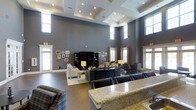 The Club Room is a single-floor communal space designed to provide an upscale, welcoming environment for gatherings and relaxation. The room's most striking architectural feature is its dramatically high ceiling, accented by a coffered tray design with recessed lighting that adds depth and dimension to the space. Clerestory windows positioned near the top of the walls allow natural light to filter in from multiple directions, complementing the warm glow of the recessed and lamp lighting throughout.

The walls are finished in a rich slate gray, providing a sophisticated backdrop that ties the space together. The layout is thoughtfully organized into distinct zones. A lounge area features a large sectional sofa and armchairs arranged around a dark entertainment center with a wall-mounted television. Adjacent to this is a more intimate seating cluster with upholstered chairs, suitable for smaller conversations. Framed artwork decorates the walls, adding a personal, curated touch.

A granite-topped bar counter is prominently positioned toward the front, offering a functional space ideal for entertaining. The polished concrete-style flooring grounds the entire room, balancing the elegance of the furnishings with a clean, modern aesthetic.