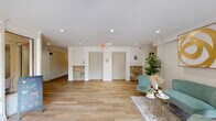 Located at 5764 Stevens Forest Rd in Columbia, Maryland, this thoughtfully designed building lobby serves as an elegant and functional entry point for residents and visitors alike. The space is anchored by warm wood-look tile flooring that extends throughout, lending a clean, contemporary aesthetic to the entire area. Near the entrance, floor-to-ceiling glass doors flood the lobby with natural light and provide a clear view of the outdoor surroundings, creating an inviting and airy atmosphere. A branded entry mat marks the welcoming threshold into the space. The seating area is tastefully furnished with teal velvet upholstered chairs and a loveseat arranged around gold-accented side tables atop a layered area rug, offering a comfortable waiting area for residents. A striking gold abstract wall art piece adds a refined decorative touch. Stacked stone accent walls flank the elevator bay, providing a rich textural contrast to the otherwise clean white walls and trim. Recessed lighting throughout ensures the space is well-lit at all times. A tall potted indoor tree adds a natural, lively element to the interior. Adjacent corridors lead residents to building amenities, including a pet wash station and additional common areas visible through interior windows, highlighting the comprehensive functionality of this well-appointed lobby.