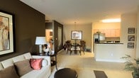 This single-floor residence showcases an open-concept layout that connects the living, dining, and kitchen areas into one cohesive, flowing space. Light-toned tile flooring extends throughout, creating a clean, unified foundation that enhances the sense of spaciousness. The living area features a neutral-toned sofa accented with decorative pillows, complemented by a sleek side table and lamp. A bold dark-painted accent wall adds depth and contrast to the otherwise warm, neutral color palette, creating a striking visual anchor.

The dining area sits centrally within the open plan, furnished with a dark wood table and matching chairs beneath an elegant chandelier that serves as a focal point. A framed mirror and decorative vase with branches add refined touches to the space.

Adjacent to the dining area, the kitchen features light wood cabinetry, dark countertops, and stainless steel appliances, including a full-size refrigerator. A peninsula-style counter provides additional workspace and subtly defines the kitchen zone without disrupting the open flow. Overall, the space balances modern design elements with warm, comfortable touches, resulting in a practical yet stylish interior.