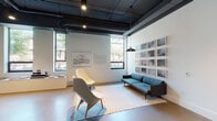 This single-floor space presents a refined blend of contemporary design and artistic curation, creating an environment that is both functional and visually engaging. The open-plan layout flows seamlessly between distinct zones, each with its own character and purpose.

The main lounge area features exposed black structural ceilings with industrial-style pendant lighting, polished concrete and warm-toned floors, and an array of curated framed artwork adorning the white walls. Modern Scandinavian-style lounge chairs and a sleek teal sofa invite relaxed gathering, while large windows frame views of the surrounding streetscape and greenery, flooding the interior with natural light.

A quieter seating nook offers a circle of upholstered chairs around a low round table beneath a minimalist pendant fixture, with floor-to-ceiling glass doors opening to views of lush outdoor plantings. Adjacent to this is a dining area with an oval table and warm-toned wood chairs, accompanied by a circular mirror and elegant pendant lighting.

The space also includes a service counter with a marble-look surface, dark wood cabinetry, and dual mounted screens — well-suited for reception or hospitality use. Large glass walls reveal an enclosed courtyard with lounge seating, greenery, and open-air gathering areas, connecting the interior to the outdoors effortlessly.