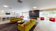 This single-floor communal space is designed with a contemporary aesthetic that balances bold accents with clean, neutral architecture. The main lounge area is anchored by a striking dark-paneled feature wall housing a sleek linear fireplace, flanked by floor-to-ceiling windows that flood the space with natural light and offer views of the surrounding landscaping. A warm wood-slat ceiling detail above this zone adds texture and visual warmth, complemented by elegant pendant lighting.

The lounge is furnished with pairs of vibrant chartreuse sofas arranged around sculptural coffee tables, while bold red accent chairs provide additional seating throughout. A dark area rug defines the central seating zone, contrasting with the light large-format tile flooring that extends across the rest of the space. A wall-mounted flat-screen television serves a secondary seating area with darker flooring and paired charcoal accent chairs.

Adjacent to the lounge is a well-appointed kitchenette area featuring a long white countertop island with black bar stools, pendant lighting overhead, warm wood cabinetry, and a sink with under-counter appliances. The overall ambiance is modern, inviting, and well-lit, making it well-suited as a shared social or gathering space.