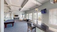 This inviting community amenity space is thoughtfully designed to accommodate a variety of social and recreational activities. The interior is finished in a calming sage-green and soft gray palette, complemented by plush blue-toned carpeting and crisp white trim throughout.

The main gathering area features soaring coffered ceilings with elegant beams, recessed lighting, and large windows fitted with blinds that frame views of the surrounding outdoor landscape. French doors provide direct access to an outdoor area, filling the space with an abundance of natural light. Comfortable seating arrangements, including upholstered armchairs and a sofa, create relaxed conversation areas, while an arcade-style gaming table adds a playful touch.

A dedicated kitchenette area is equipped with white cabinetry, dark granite countertops, stainless steel appliances including a refrigerator and microwave, and a bar-height counter with seating for casual dining. Pendant globe lights add a stylish finishing touch overhead.

A separate game room features a pool table, shuffleboard table, wall-mounted flat-screen television, and decorative driftwood wall art. An additional smaller room provides a quieter setting with a game table surrounded by lattice-back chairs, a window with garden views, and playful décor. Together, these spaces offer a versatile and well-curated environment for leisure and community engagement.