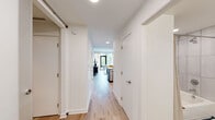 This contemporary single-floor residence showcases a clean, modern design throughout. A straight hallway with light wood flooring and crisp white walls connects the main living areas, providing an organized flow between spaces. The kitchen features a galley-style layout equipped with stainless steel appliances, dark wood cabinetry, white countertops, and a bold blue accent wall, complemented by statement pendant lighting overhead. The open living area is bathed in natural light from floor-to-ceiling windows and a glass door that frames outdoor neighborhood views, creating a bright and airy atmosphere. The primary bedroom continues the theme of generous natural light, featuring a large window with street views, light wood flooring, and a serene color palette. A second bedroom offers a cozy, well-defined space with a taupe accent wall and practical built-in shelving. Both bathrooms are finished with white tile flooring, dark wood vanities, white countertops, and large mirrors. The primary bathroom includes a walk-in closet and a glass-enclosed shower. Throughout the residence, light hardwood flooring, recessed lighting, and a neutral palette create a cohesive and contemporary living environment.