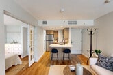 This thoughtfully designed one-bedroom, one-bathroom residence in Tewksbury, MA, makes excellent use of its compact footprint through an open-concept floor plan that seamlessly connects the living, dining, and kitchen areas. The kitchen is a standout feature, outfitted with warm wood-toned cabinetry, quartz countertops, stainless steel appliances including a refrigerator, range, and microwave, and a peninsula with bar seating that doubles as a dining space. Hardwood flooring runs throughout the main living areas, lending warmth and continuity to the space. The living room is bathed in natural light from a large window fitted with blinds and features a wall-mounted flat-screen TV with a dedicated built-in ledge. French-style glass doors connect the living area to the bedroom, which is appointed with soft sage-green walls, hardwood floors, a wide window with vertical blinds, and a generous closet. The bathroom offers a full tub-and-shower combination with tile surround, a vanity with granite countertop, and tile flooring. Neutral tones, clean lines, and modern finishes throughout create a cohesive and comfortable living environment.